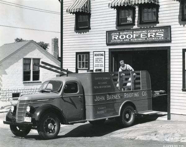 View from street towards a man at the John Barnes Roofing Company loading lumber onto the bed of an International D-30 truck parked in front of a company building. The text on the truck advertises: "Asbestos Sidewalls, Accurate Metal Weather Strips."