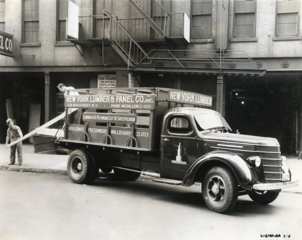 Two men load pieces of lumber onto the bed of an International D-40 truck owned by New York Lumber & Panel Company.