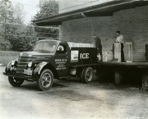Two men load blocks of ice from a loading dock onto the bed of an International D-30 truck owned by the Hygeia Ice Company.