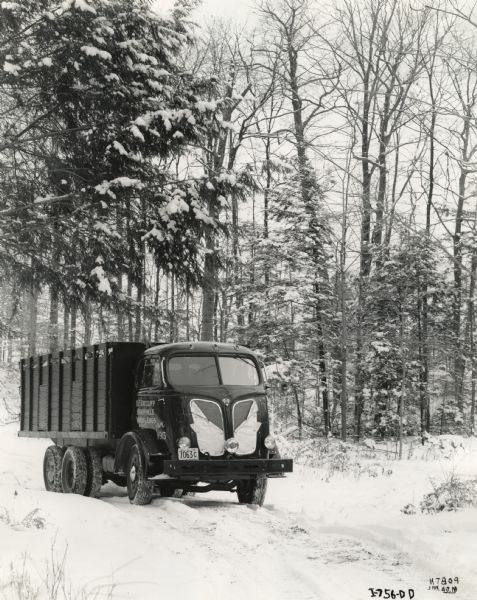 An International D-500-F truck owned by H.F. Ratcliff Wood & Logs travels along a snow-covered road through a wooded area.