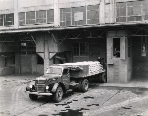 Elevated view of a man loading burlap bags from a loading dock onto the bed of an International D-50 truck owned by the Materials Transit Company.