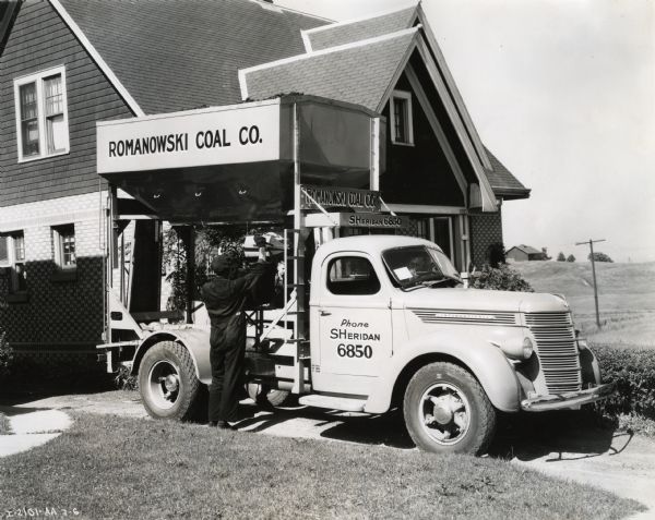 A man loads a piece of equipment onto the back of an International D-40 truck owned by the Romanowski Coal Company while it is parked in a residential driveway. A young boy is standing behind the truck.