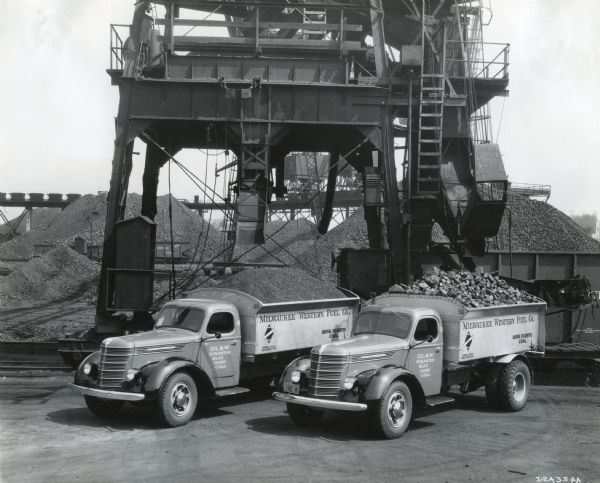 Elevated view of two International D-60 trucks owned by the Milwaukee Western Fuel Company parked near a yard with piles of coal. A large structure towering above them has chutes for filling the back of the trucks.