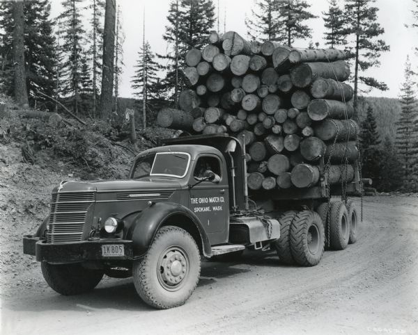 A man drives an International D-Line truck owned by The Ohio Match Company through a wooded area along a dirt road. The truck bed is laden with cut logs.