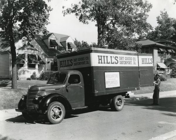 View from street of a uniformed man standing near the back of an International D-30 truck owned by Hill's Dry Goods Company parked in a residential area. The sign on the truck reads: "Headquarters for All Major Appliances. Buy on Easy Terms."