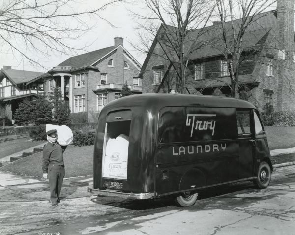 View across street towards a man loading bags of laundry into the back of an International D-Line Metro truck owned by Troy Laundry parked along the curb in a residential neighborhood. There is slush along the curb.