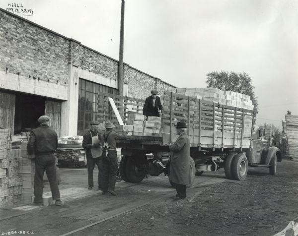 A group of men load bundles of lumber onto the bed of an International D-30 truck owned by the Kawartha Lumber Company. The truck is parked in front of a brick storage facility. A man in a long overcoat and wearing a hat stands near the truck writing on a piece of paper.
