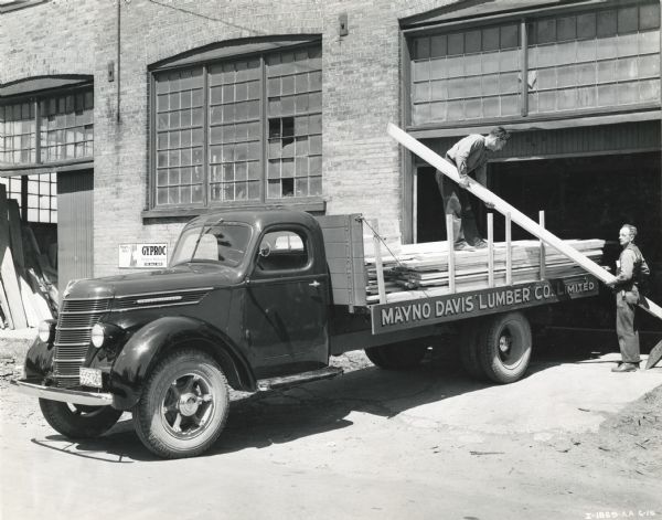 Two men load lumber onto the bed of an International D-35 truck owned by the Mayno Davis Lumber Company Limited. The truck is parked in front of what appears to be a the door of a garage or storage facility.