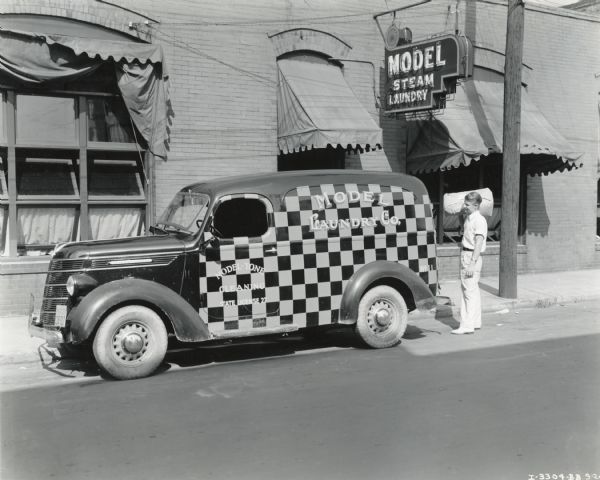 View from across street of a man loading a bag of laundry into the back of an International D-2 panel truck painted in a checkerboard pattern. The truck is parked outside the laundromat storefront.