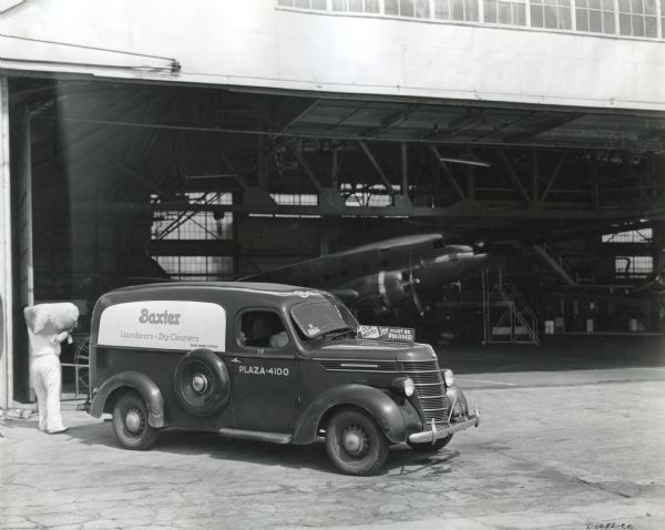 A man unloads bags of laundry from the back of an International D-2 panel truck owned by Baxter Laundry. The truck is parked near a hangar and inside is an airplane.