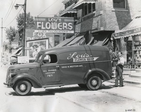 View from street of man loading a bouquet into the back of an International D-2 panel van owned by Lou's Flower Shop. The truck is parked along a commercial street in front of Lou's storefront.