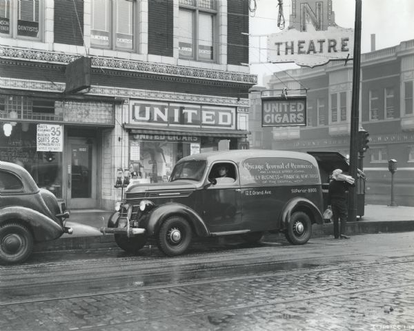 View from across street of a man unloading newspapers from the back of an International D-Series panel truck owned by the Charles Levy Circulation Company. A man is sitting in the driver's seat, and the text on the side of the truck reads: "Chicago Journal of Commerce and La Salle Street Journal. The Daily Business and Financial Newspaper of the Central West. 12 E. Grand Ave. SUPerior-8900."