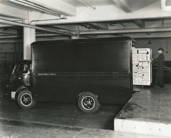 A man unloads a rolling cart filled with wrapped packages from an International D-300 truck onto a loading dock. The truck was used by United Parcel Service to collect packages from department stores.