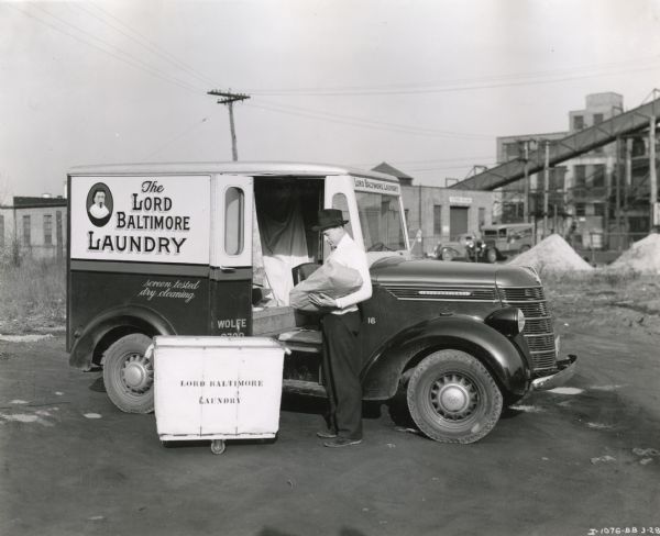 A man unloads packages from an International D-2 truck owned by Lord Baltimore Laundry. A wheeled laundry basket stands near the truck's opened side door.