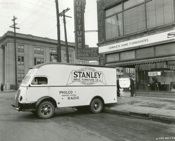 Two men loading what appears to be a cabinet into the back of an International D-300 truck owned by the Stanley Brothers Furniture Company. The truck is parked in front of the company's storefront near an intersection in a commercial area.