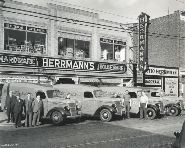 View across street of men posing beside a fleet of International trucks owned by the Otto Herrmann Hardware Company. The trucks are pared outside the company's store at 6729 Myrtle Avenue.