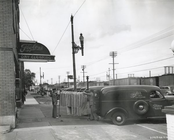 A fleet of International Model D-2 trucks are parked along the curb outside Figler's Dry Cleaning. Two men unload a rack of clothing in garment bags from the truck in the foreground. In the background are railroad cars.