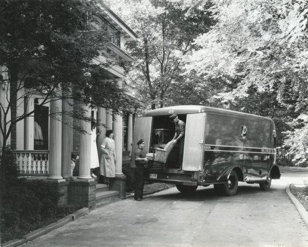 Two uniformed men loading or unloading an upholstered chair from the back of an International C-300 truck parked in front of a residence. Two women and a dog are looking on from the home's porch.