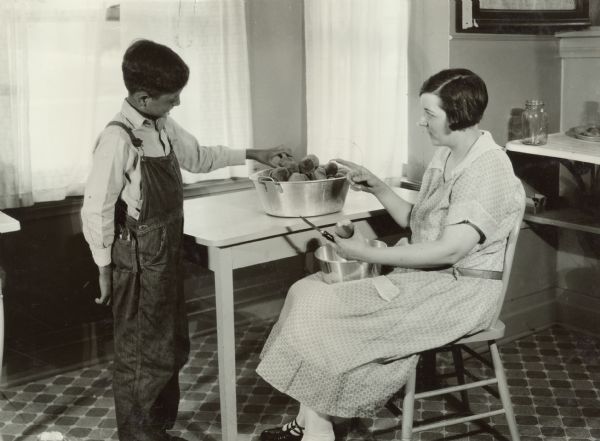 A woman preparing peaches for canning. She appears to be shaking her finger at a young boy as he reaches for a peach. This scene occurred on the Harvester Farm.