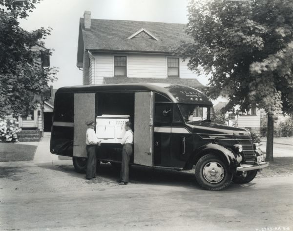 Two men unload what appears to be an oven from an International D-35 truck owned by the Sibley, Lindsay, and Curr Company department stores. The truck is parked on a dirt road in front of a residence.