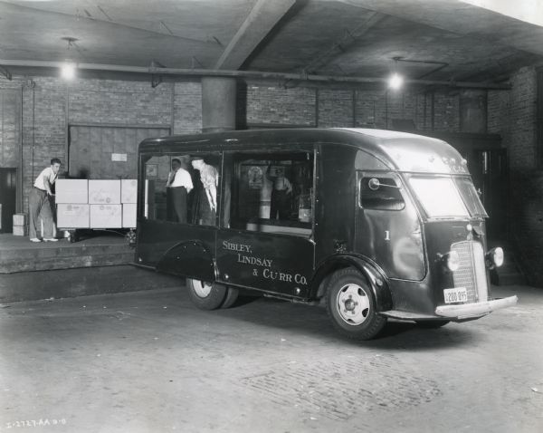 Men place boxes from a rolling cart on a loading dock onto an International C-300 truck owned by the Sibley, Lindsay, and Curr Company department store. One man stands inside the truck which has large windows on both sides.