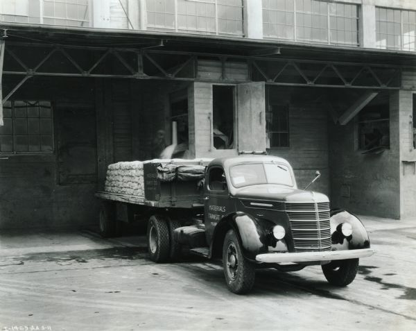 An International D-50 truck carrying fire-building materials parked near a loading dock. A man is standing near the back of the truck loading one of the sacks.