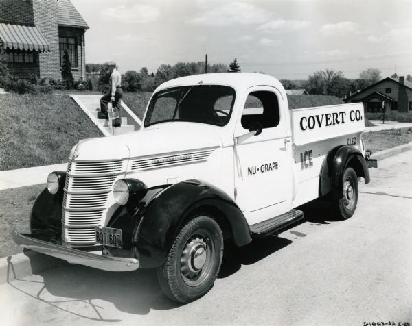 A man climbs the steps to a residence as he delivers a block of ice. An International D-2 truck owned by the Covert Company parks along the curb. The text on the truck reads: "Nu-Grape. Covert Co. Ice. Cleo."