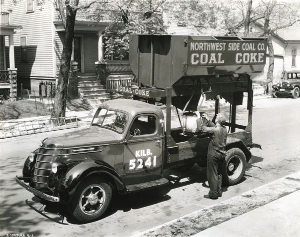 Elevated view of a man filling a bucket with coal from a dispenser on the back of an International truck owned by Northwest Side Coal Company. The text on the truck reads: "Kilb. 5241" and advertises "Coal - Coke."