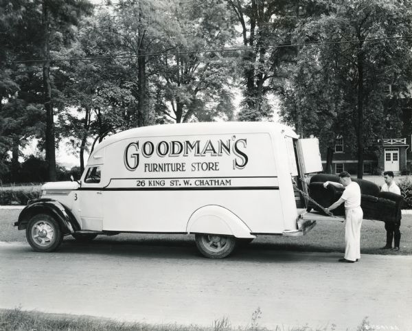 Two men unloading a sofa from the back of an International D-30 truck owned by Goodman's Furniture Store. The truck is parked in a residential neighborhood.