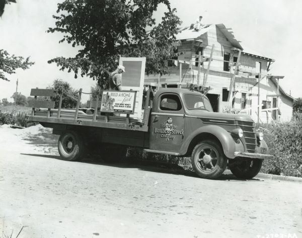 A man unloads a door from the bed of an International D-35 truck owned by Fort Wayne Builders Supply Company. A house under construction is in the background.