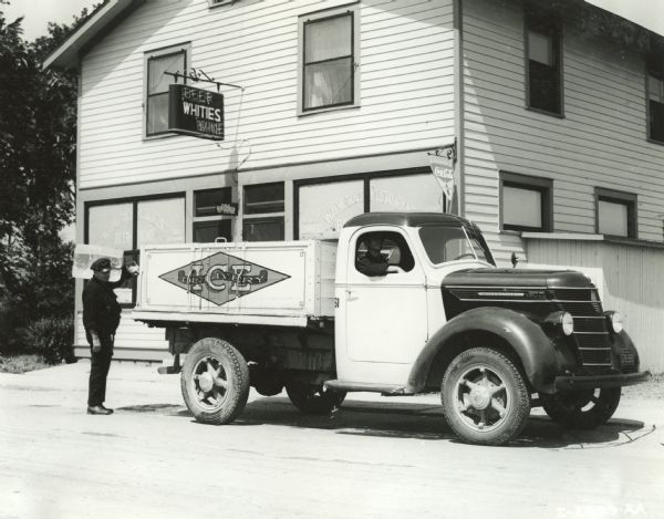 A man unloads a block of ice from the back of an International D-30 truck owned by the Ice Delivery Company. The truck is parked in front of a restaurant.