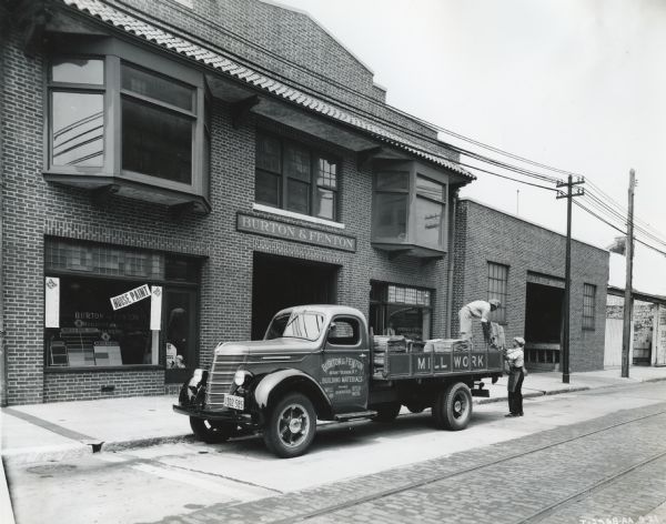 Men load what appears to be lumber onto the bed of an International truck owned by Burton & Fenton Building Materials. The truck is parked in front of the Burton & Fenton storefront.