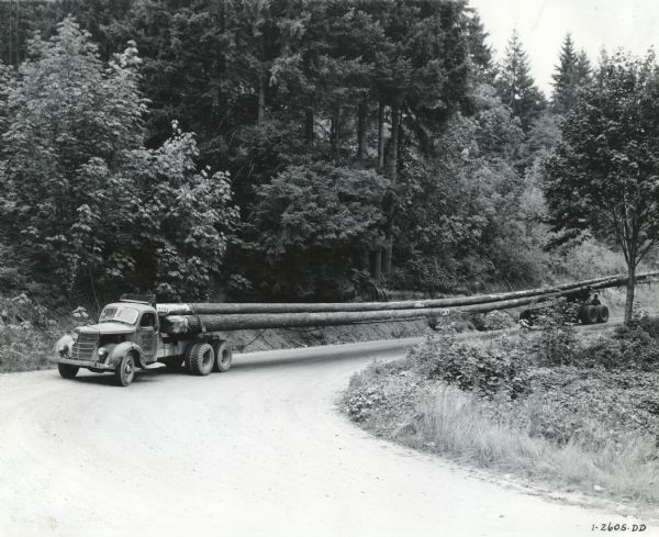 An International D-line truck carries timber down a dirt road in a wooded area. The logs are so long, they require two trucks to carry them. The text on the door of the truck reads, "WISCRAIC. 34000. Gross."