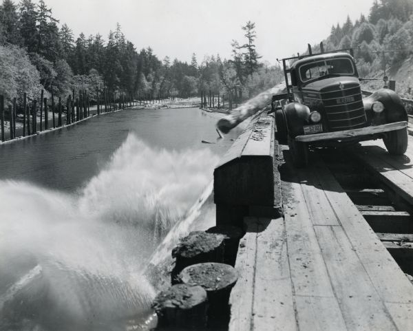 An International truck is dumping logs into the North River while positioned on a wooden platform high above the bank.