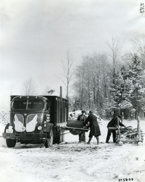 Three men load a pile of logs onto a trailer attached to an International truck in Ontario, Canada. There appears to be a belt-driven saw attached to the side of the truck for cutting the logs before loading. A snow-covered forest is in the background.