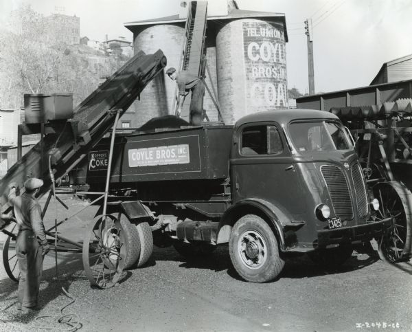 A man stands on the bed of an International DR-500 truck while shoveling coal onto a conveyor operated by another man on the ground. The truck was owned by Coyle Brothers Incorporated, purveyors of coal and coke. Storage silos are in the background.