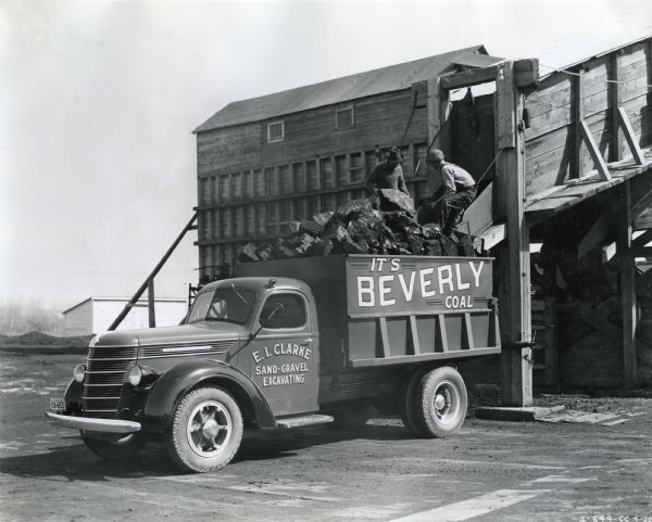 Men loading large chunks of coal onto the bed of an International D5-40 truck owned by E.I. Clarke Sand and Gravel Company. A storage building is in the background.