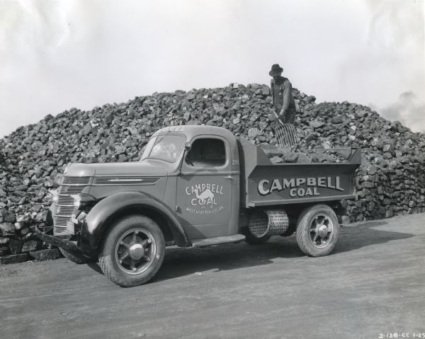 A man standing on a large pile of coal uses a rake to load coal onto the bed of an International D-30 truck owned by Campbell Coal. The driver's side door features an illustration of a camel and the text: "Campbell Coal 'Most Heat Per Dollar.'"