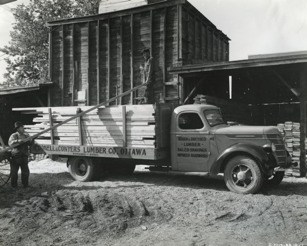 Two men load a piece of lumber onto the bed of an International D-35 truck owned by McDonell and Conyers Lumber Company of Ottawa, Ontario, Canada. Additional boards are piled beneath a shed in the background.