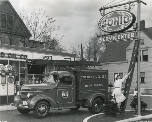 An employee of Sohio's Neon Patrol uses a ladder to reach a neon sign to make repairs. An International D-15 truck owned by Sohio is parked in the driveway beside him, and the service station and gasoline pumps are in the background. The text on the side of the truck reads: "Sohio X-70. Standard Oil Co., Ohio. Neon Patrol." Sohio was also known as Standard Oil of Ohio.