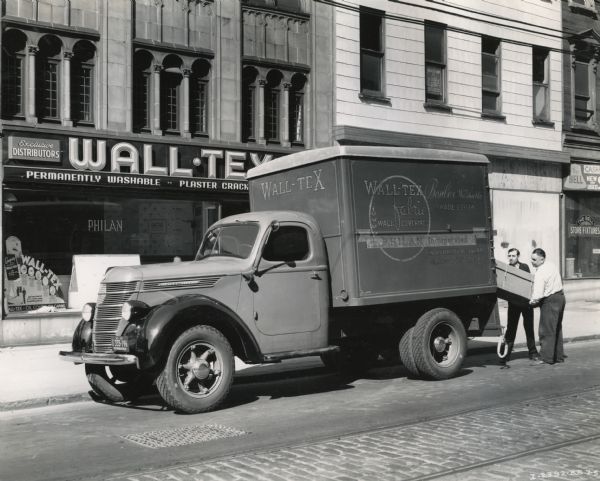 Two men load a box into the back of an International D-35 truck owned by Philan, Incorporated, a distributor of fabric wall coverings. The company storefront is behind the truck.