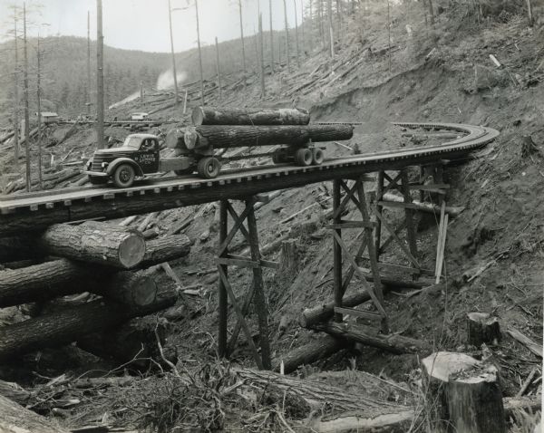 An International DR-60 motor truck owned by the Lewis Lumber Company hauls a load of logs over a wooden bridge spanning cutover land.