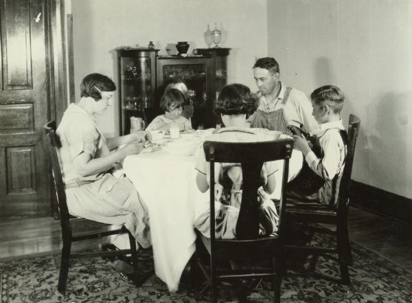 Farm family seated at table eating a meal at the Harvester Farm. Original caption reads: "Comparison picture to show close-up of canned foods served."