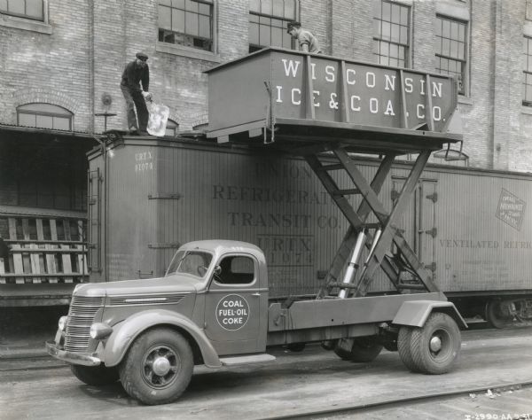 A man stands in a lift attached to the bed of an International Model DR-60 truck owned by the Wisconsin Ice and Coal Company. Another man stands atop a railroad car and unloads a block of ice using metal tongs. There is a large brick building behind them with a loading dock.