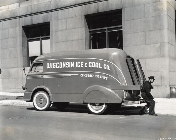 View from street of man carrying ice out of the rear bumper of an International D-300 truck owned by the Wisconsin Ice & Coal Company. The text on the truck advertises "Ice Cubes — Ice Chips."