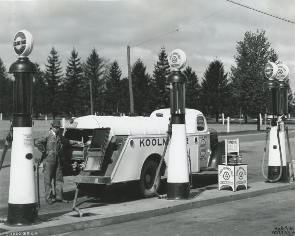 A man stands near the back of an International D-30 truck owned by Cowie Brothers City Service Company as the truck supplies a service station with oil. The truck is parked near a set of gasoline pumps.
