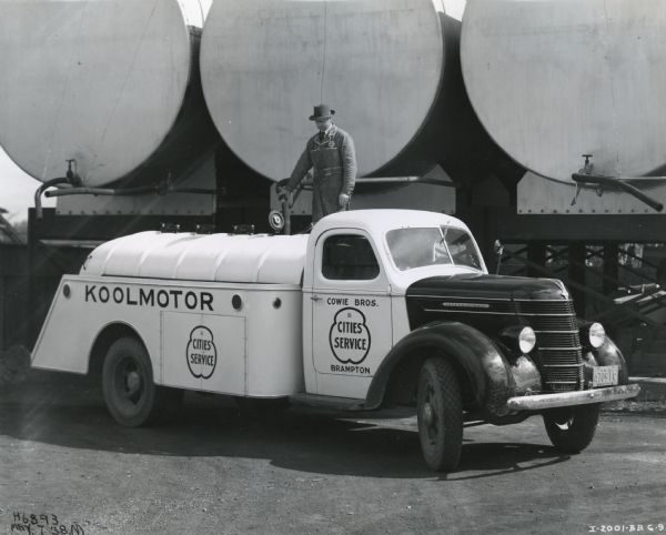A man stands atop a platform behind an International D-30 truck as he fills it with oil from a tank. The truck was outfitted with a 500-gallon 5-compartment tank and owned by Cowie Brothers Cities Service.