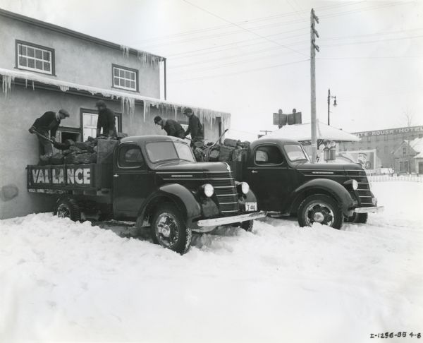 Men use shovels and pitchforks to unload large pieces of coal from the beds of two International D-30 trucks into a small doorway of a building. The trucks are owned by the Vallance Cartage and Fuel Company. The White House Hotel, located at 346 Second Avenue South, is behind the trucks, and snow is on the ground. Icicles are hanging from the eaves of the building.