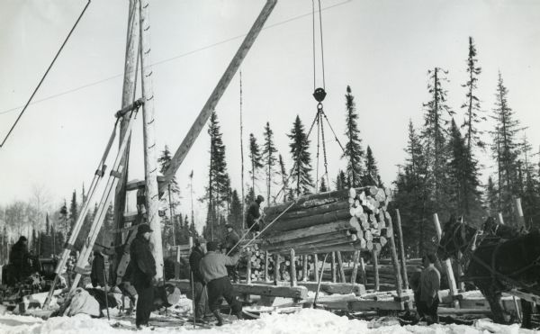 A group of men from the Newaygo Lumber Company use a pulley system to lift logs off the snow-covered ground. A team of horses stands on the right.