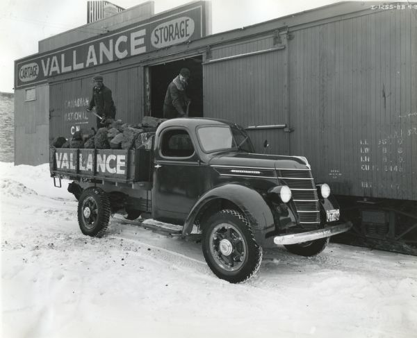 Two men load large pieces of coal onto the bed of an International D-30 truck from the open door of a railroad boxcar. There is a storage building in the background.